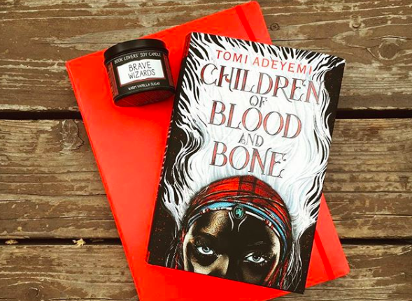 Novel CHILDREN OF BLOOD AND BONE on top of red journal next to "brave wizards" candle