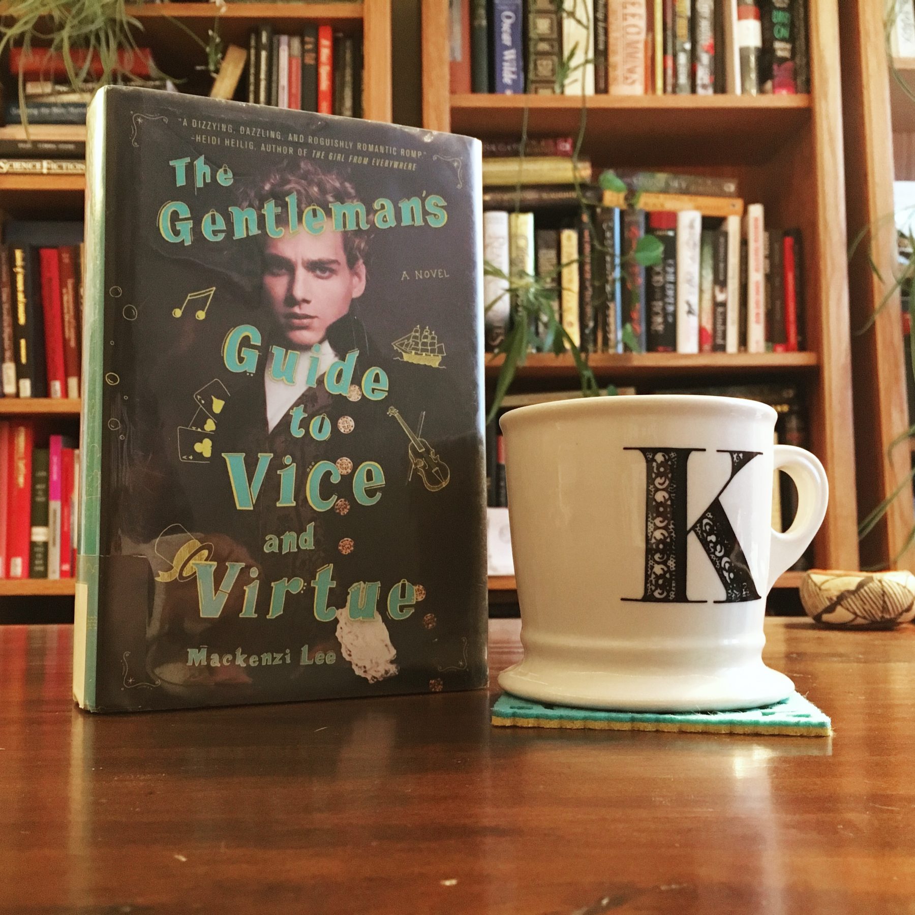 Hardcover copy of Mackenzi Lee's A Gentleman's Guide to Vice and Virtue sits on a dark wood desk next to a white mug with a "k" on it. Bookshelves in the backgroud