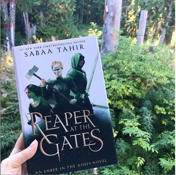 White hand holds A REAPER AT THE GATES with lush forest in the background