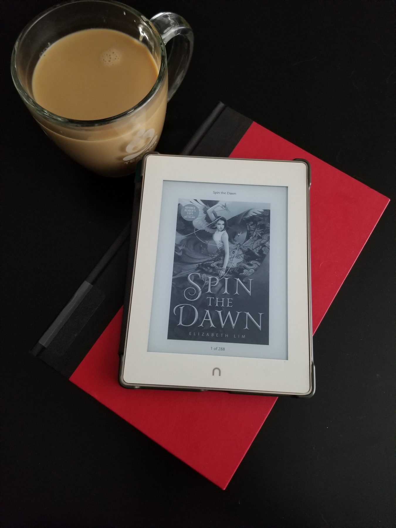 Ereader with SPIN THE DAWN on top of a red book next to a cup of coffee