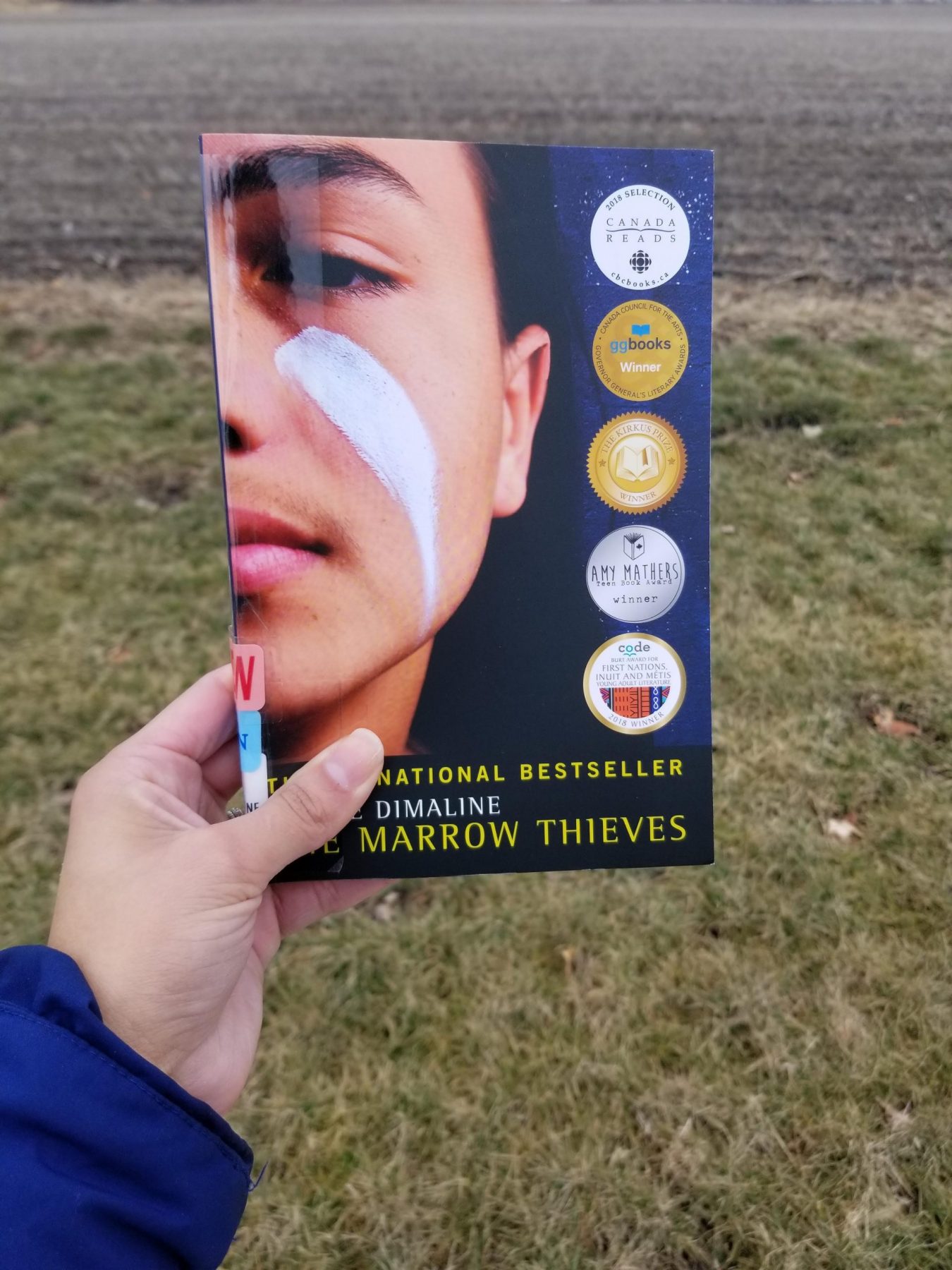 Paperback copy of The Marrow Thieves held in J's left hand. A natural landscape of greens and browns is in the background.