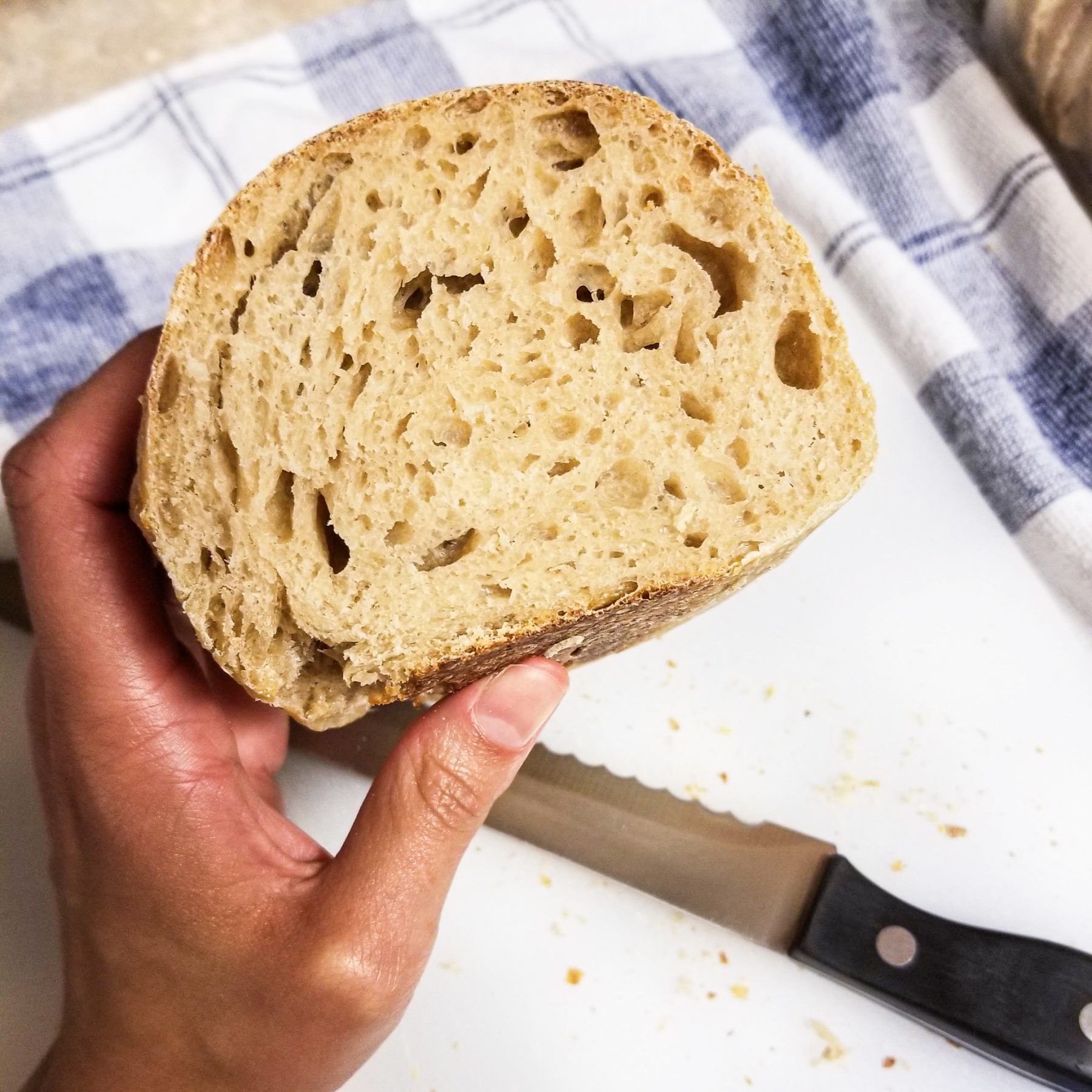 A light-skinned Black person holds a slice of sourdough in her left hand. A cutting board, blue and white gingham cloth and serrated knife lie below.