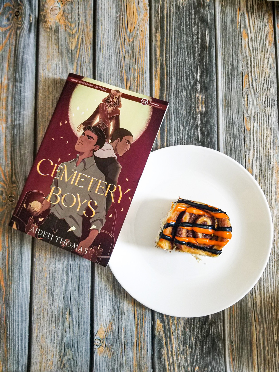 Novel CEMETARY BOYS by Aidan Thomas rests on a wood plank background. A white plate sits beside the book with a cinnamon roll that has black and orange striped icing