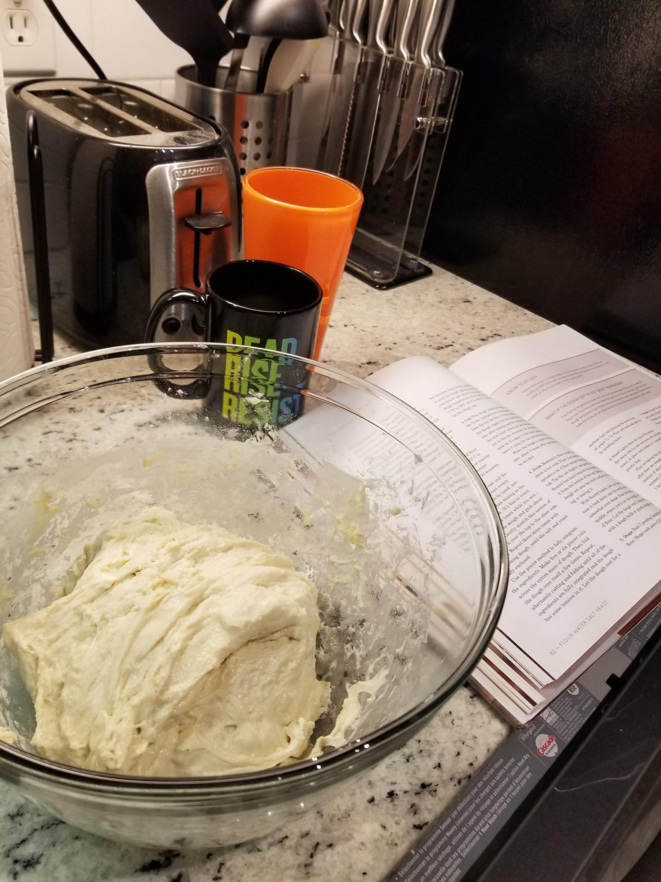 a glass bowl of dough on a marble counter next to an open recipe book