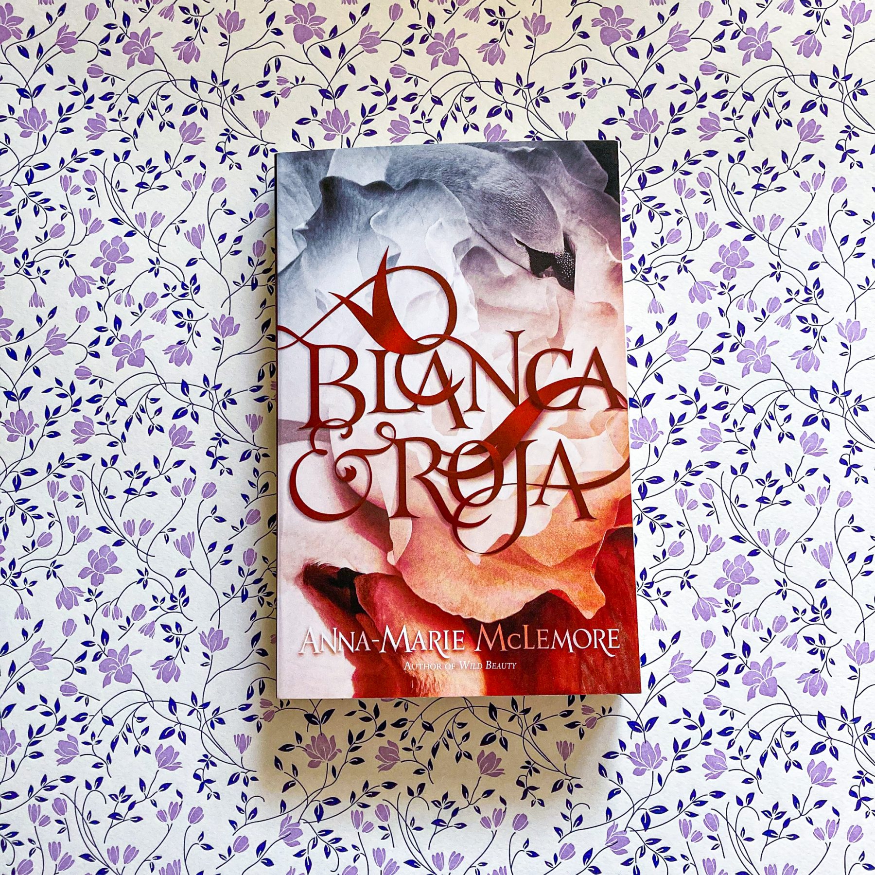 Paperback copy of Blanca & Roja rests on background of purple and navy flowers