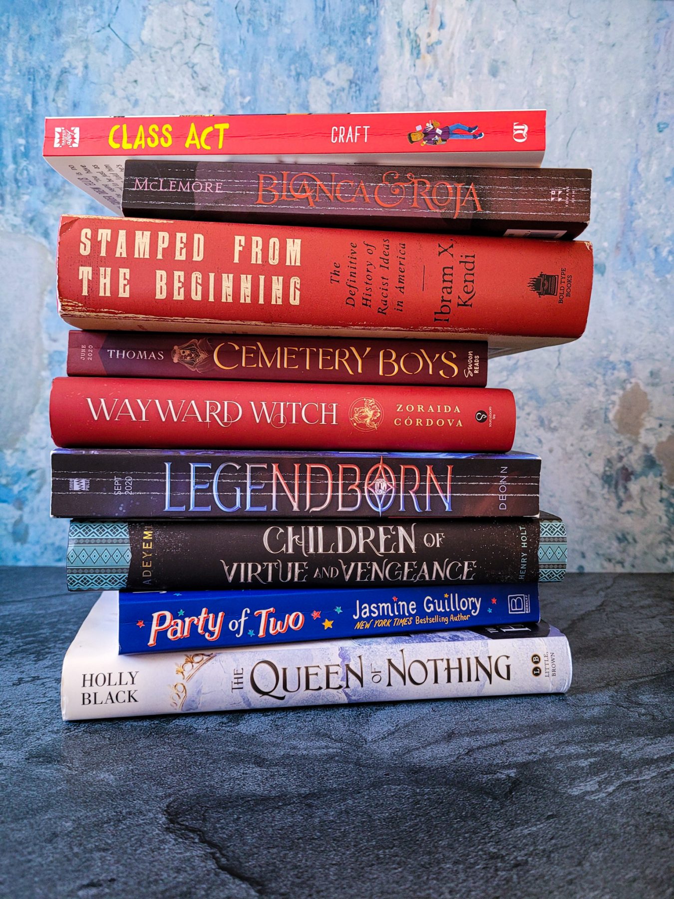 a stack of books sits in front of a blue and black marble background. From top to bottom: Class Act by Jerry Craft, Blanca & Roja by Anna-Marie McLemore, Stamped from the Beginning by Ibram X Kendi, Cemetery Boys by Aiden Thomas, Legendborn by Tracey Deon, Children of Virtue and Vengeance by Tomi Adeyemi, Party of Two by Jasmine Guillory and Queen of Nothing by Holly Black