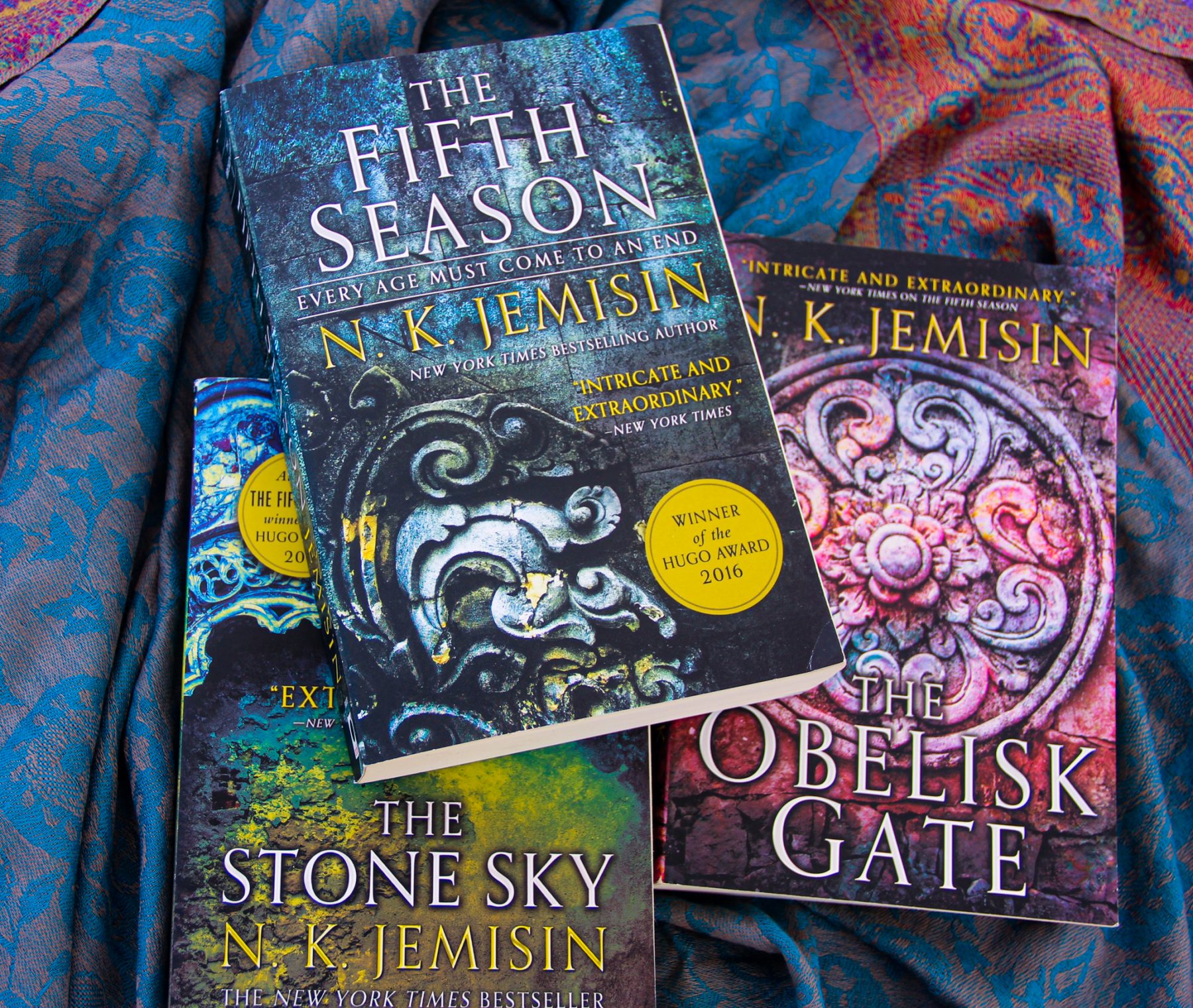 Paperback copies of The Fifth Season, The Stone Sky and The Obelisk Gate stacked on blue fabric