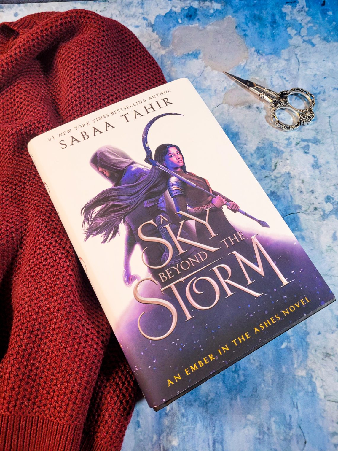 Book, A Sky Beyond the Storm on a blue and grey marble background with a red sweater underneath the book on the left, and a pair of vintage scissors on the right