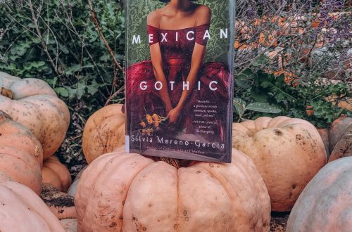 Mexican gothic sits atop a pile of orange pumpkins with scrub brush in the background