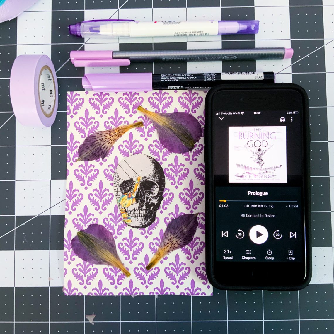 light purple stationary items, pens, Washi tape beside an iPhone with the screen showing The Burning God on audiobook