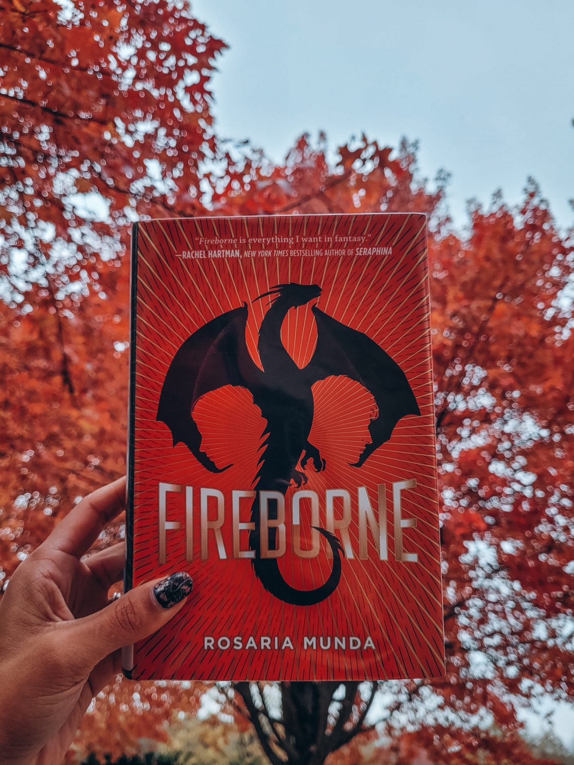 Fireborne held aloft by a light brown hand with black nail polish. bright red leaves and trees in the background
