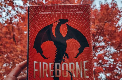 Fireborne held aloft by a light brown hand with black nail polish. bright red leaves and trees in the background