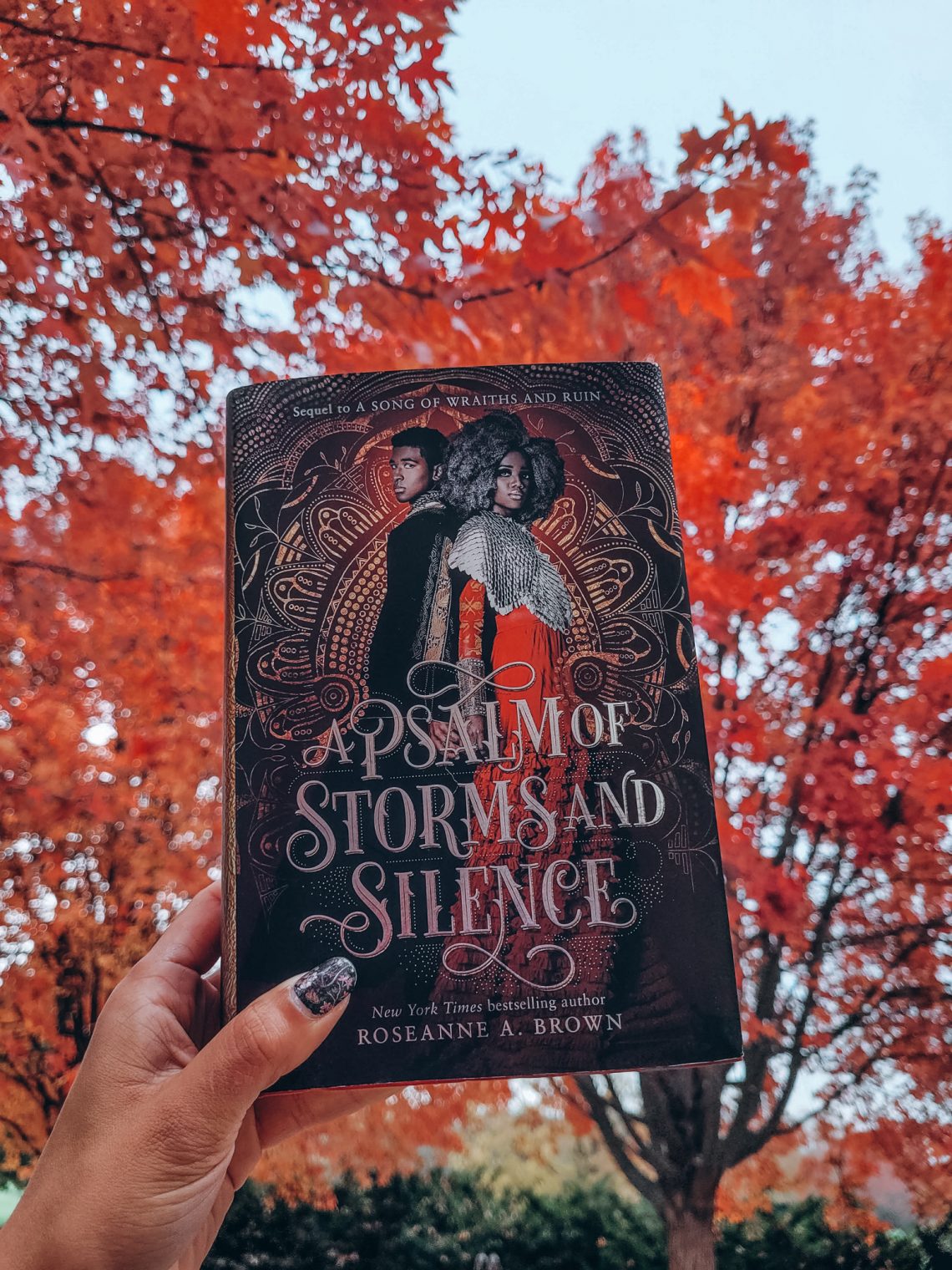 A light brown hand with dark nail polish holds aloft the hardcover of A Psalm of Storms and Silence. Trees with bright red autumn leaves and a cloudy sky form the background