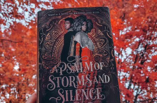 A light brown hand with dark nail polish holds aloft the hardcover of A Psalm of Storms and Silence. Trees with bright red autumn leaves and a cloudy sky form the background