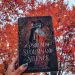 A light brown hand with dark nail polish holds aloft the hardcover of A Psalm of Storms and Silence. Trees with bright red autumn leaves and a cloudy sky form the background