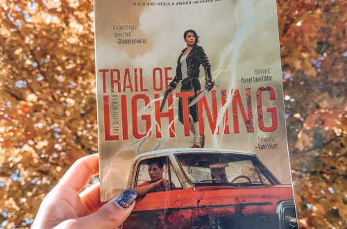 Trail of Lightning held by a light brown hand with black nail polish. gold autumn leaves in the background