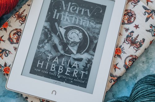 Merry Inkmas on white e-reader on top of christmas wrapping paper and between skeins of yarn