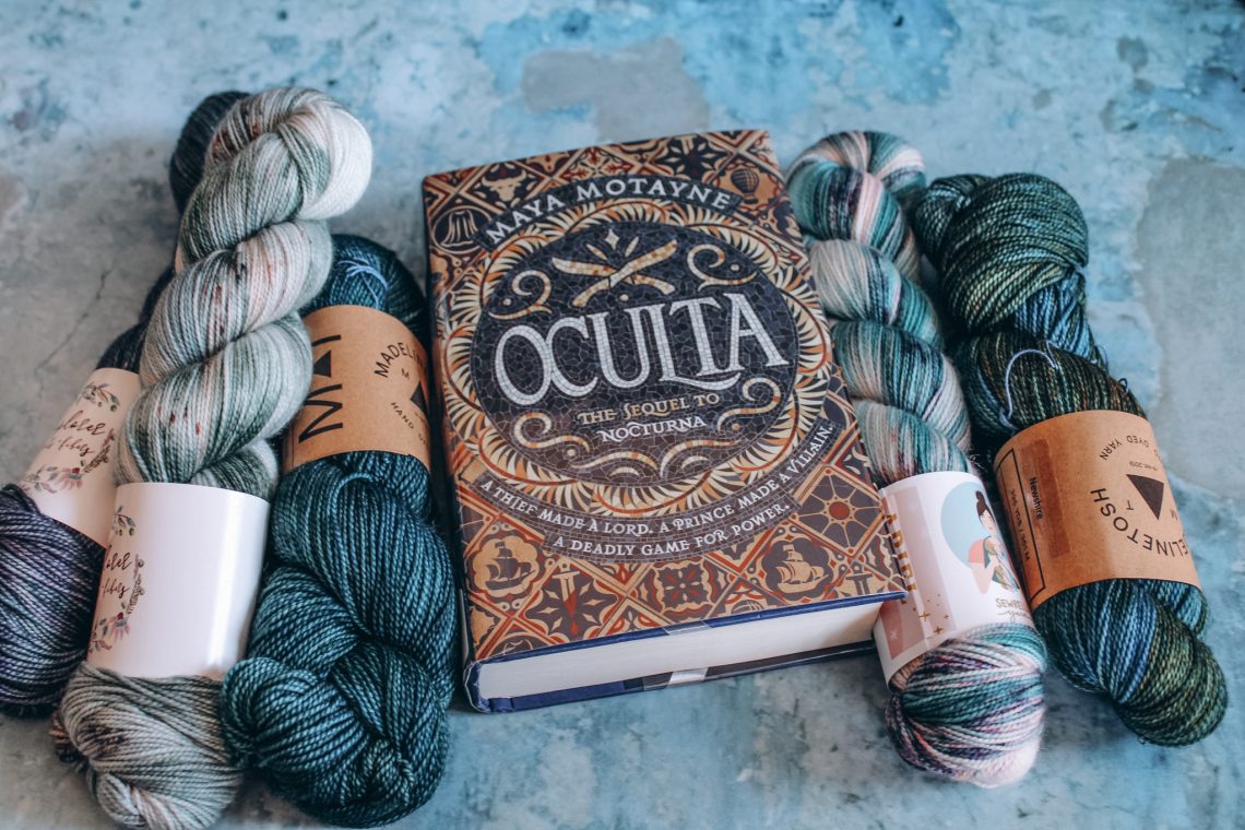 Oculta on a blue marble background, flanked by skeins of blue, teal and white yarn