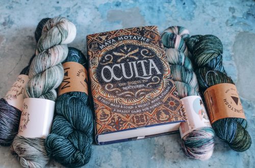 Oculta on a blue marble background, flanked by skeins of blue, teal and white yarn