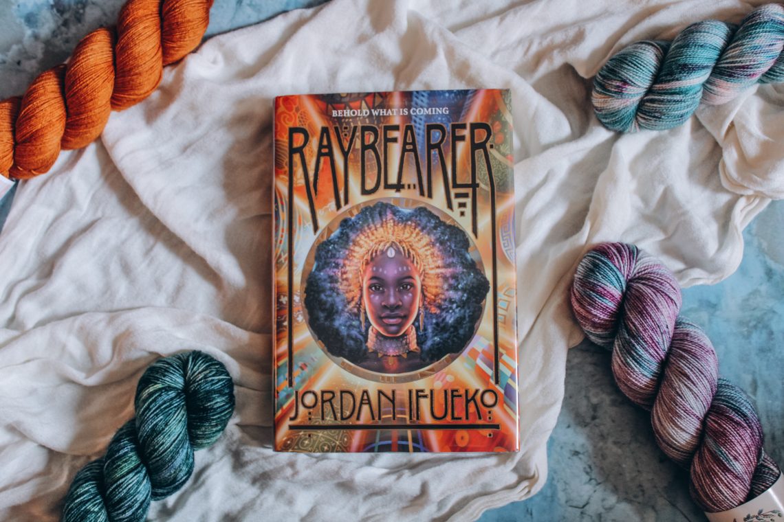 Hardcover copy of Raybearer on a white cloth, surrounded by 4 skeins of jewel-toned yarn on each corner