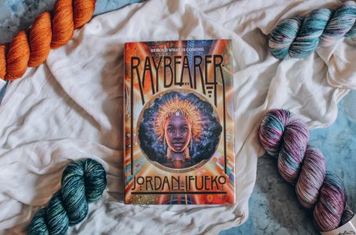 Hardcover copy of Raybearer on a white cloth, surrounded by 4 skeins of jewel-toned yarn on each corner