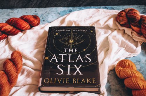 The Atlas Six on a white cloth and blue marble background. 4 skeins of orange yarn flank the book on each corner