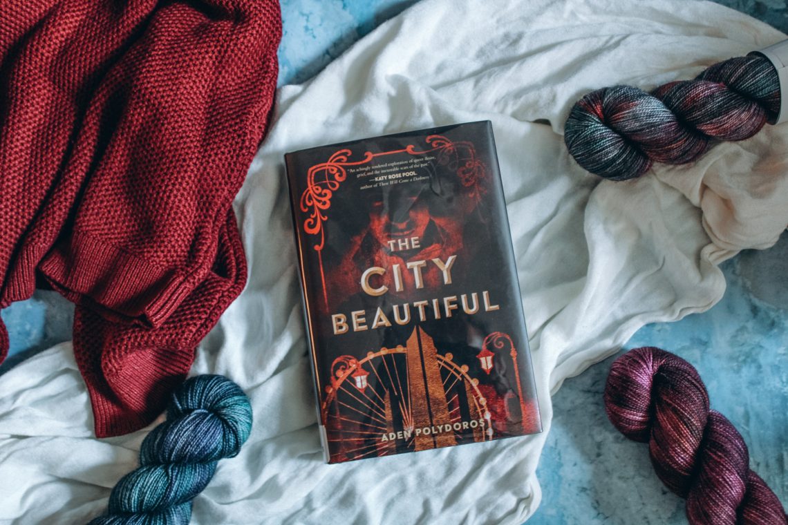 Library copy of The City Beautiful on a white cloth, three skeins of yarn surround it and a red sweater lays to the left