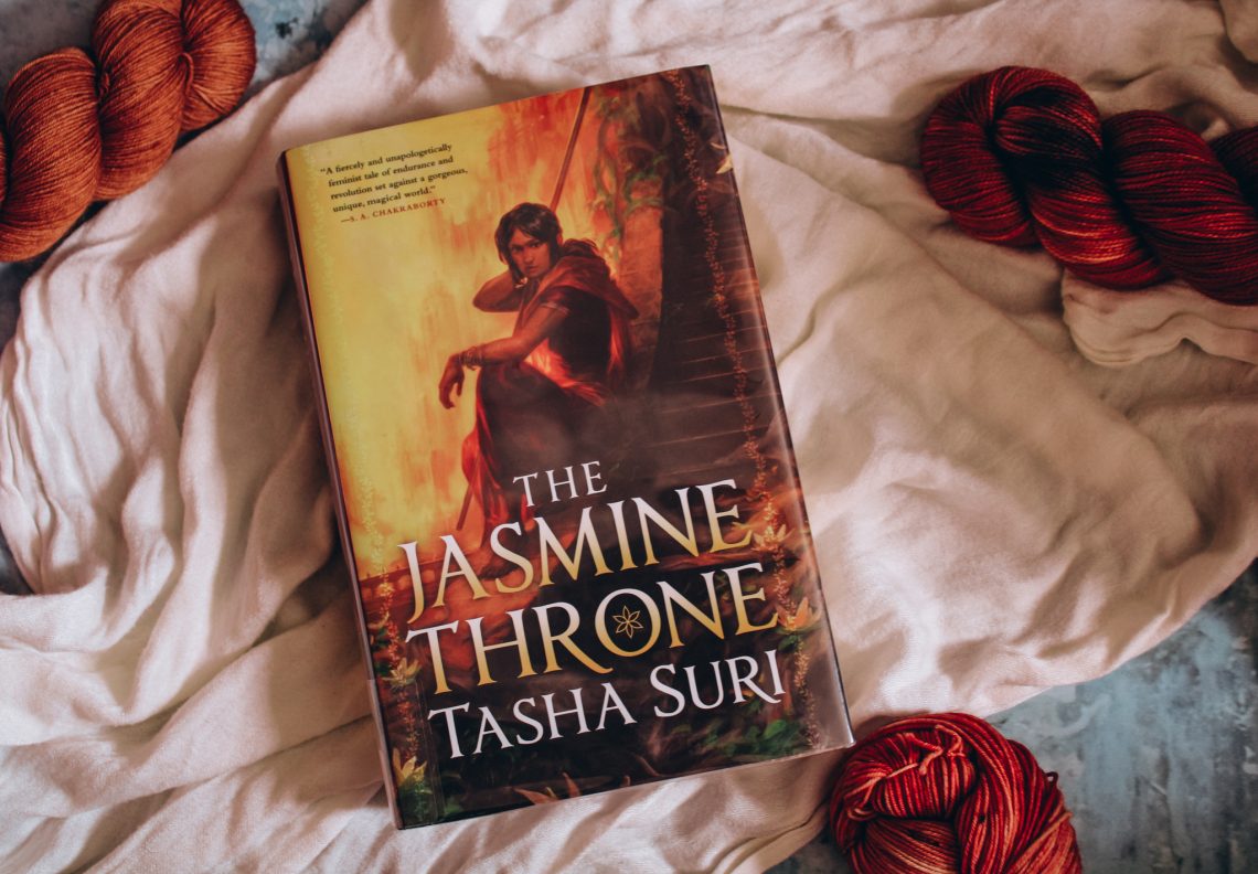 Hardcover of The Jasmine Throne on top of a light cloth and surrounded by 4 bright orange skeins of yarn