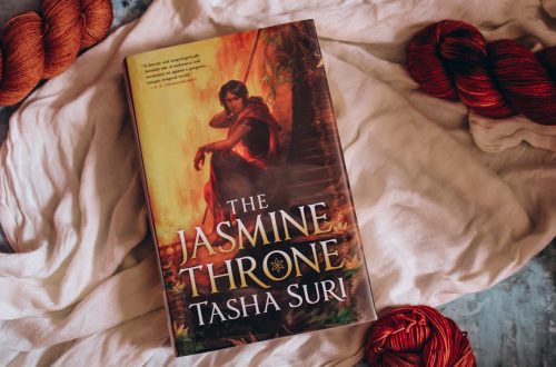Hardcover of The Jasmine Throne on top of a light cloth and surrounded by 4 bright orange skeins of yarn