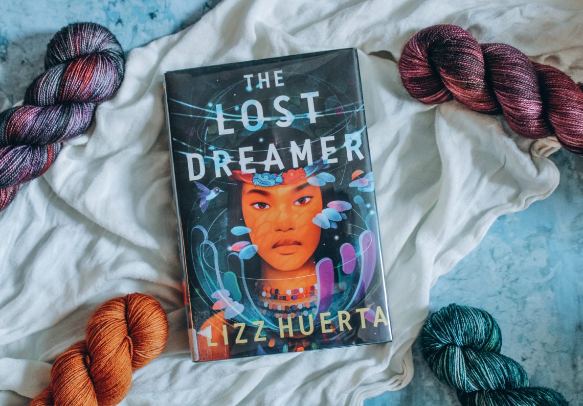 The Lost Dreamer surrounded by 4 skeins of grey, maroon, gold, and teal yarn on a white cloth and blue marble background.