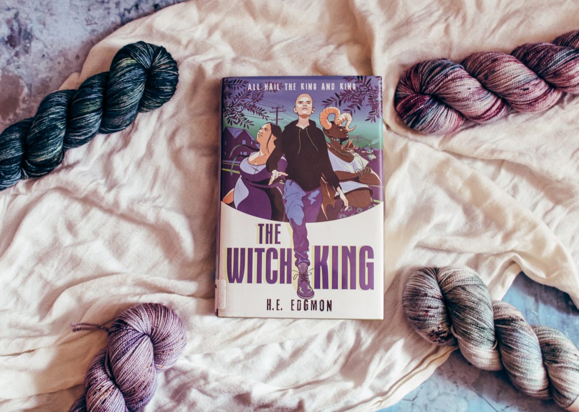 The Witch King on a white cloth surrounded by 4 blue, purple, and cream colored yarn skeins