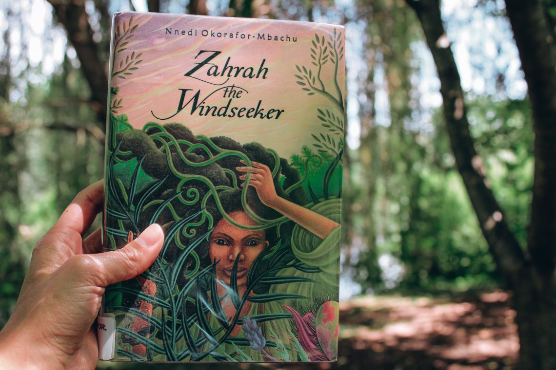 A light brown hand holds Zahrah the Windseeker in front of a lush forest