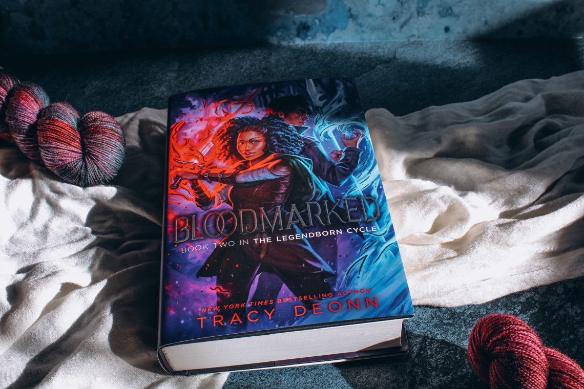 light streaks across a copy of Bloodmarked by Tracy Deonn on top of a white cloth and blue marble background