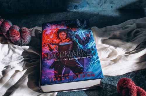 light streaks across a copy of Bloodmarked by Tracy Deonn on top of a white cloth and blue marble background