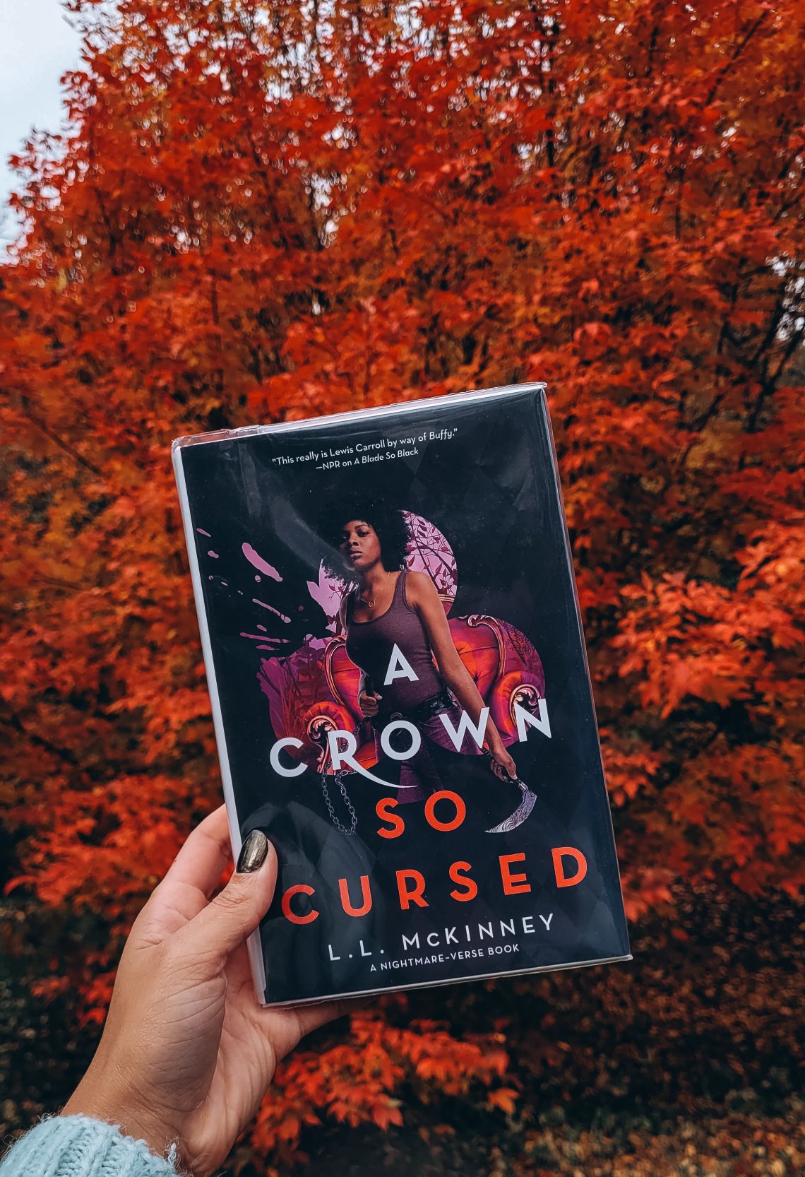 A light brown hand holds A Crown So Cursed in front of a blazing red tree.