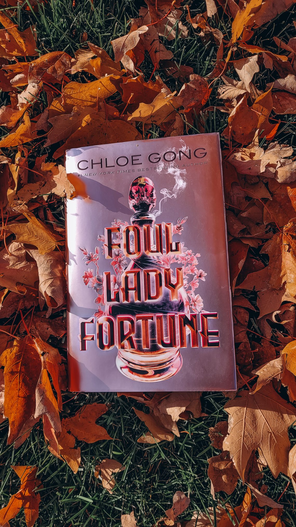 A pink metallic hardcover copy of Foul Lady Fortune rests on yellow and orange fallen leaves.