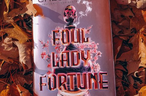 A pink metallic hardcover copy of Foul Lady Fortune rests on yellow and orange fallen leaves.