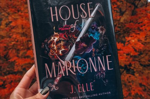 A light brown hand with black nail polish holds up a library copy of House of Marionne in front of a blazing red tree