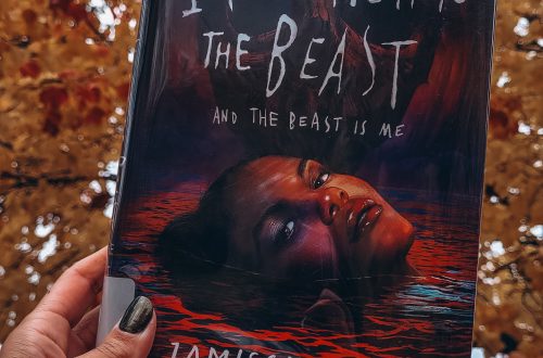 A light brown hand with black nail polish holds I Feed Her to the Beast and the Beast Is Me in front of a large tree with orange and yellow leaves.