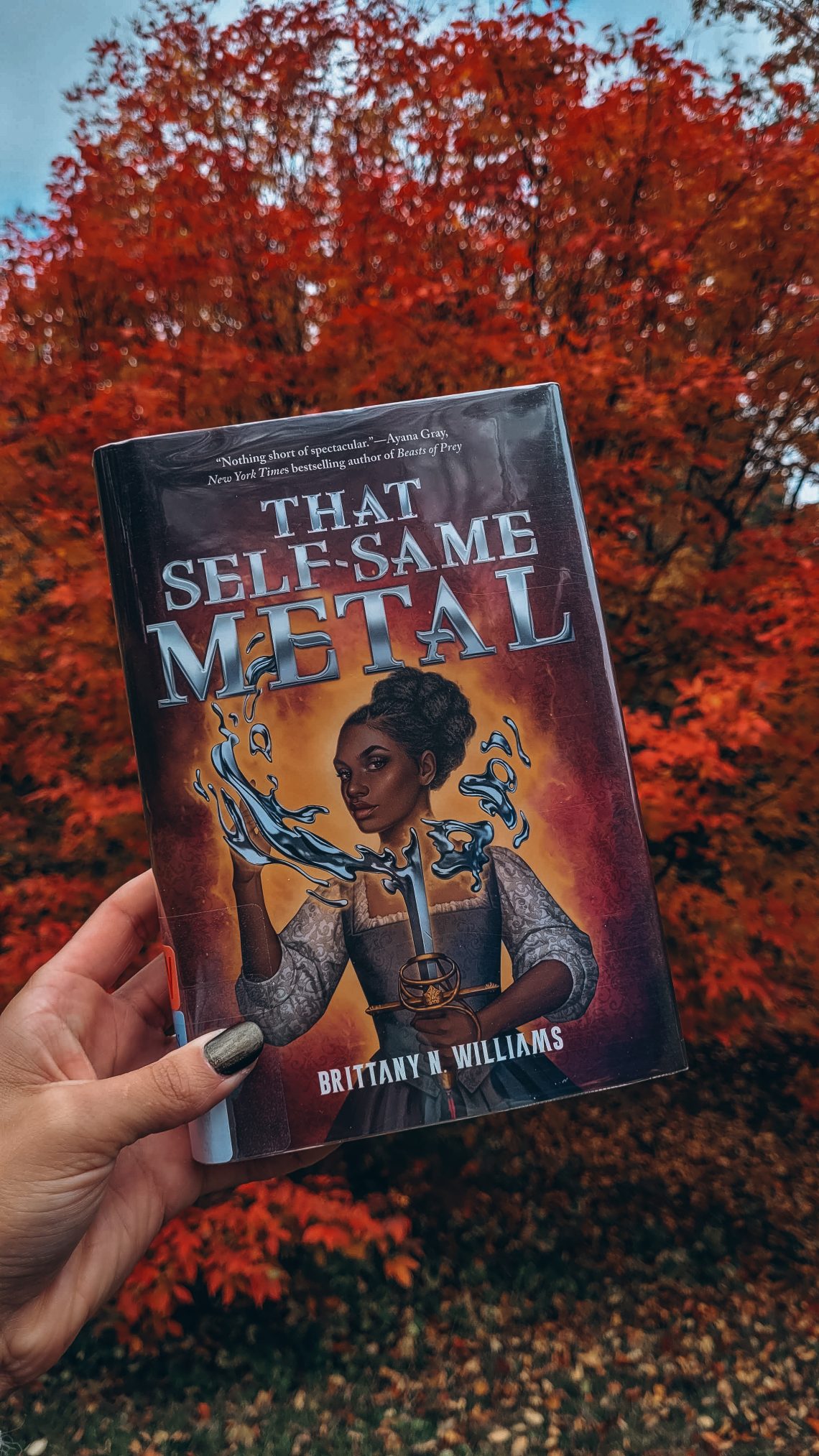 That Self-Same Metal held aloft by a light brown hand with black nail polish in front of a tree with blazing red autumn foliage.