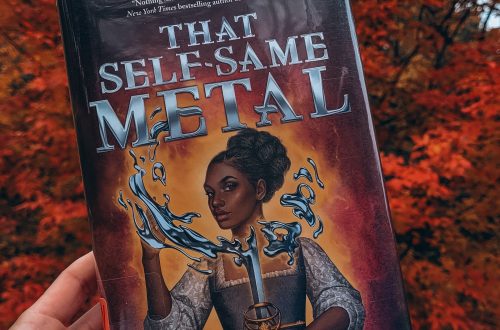 That Self-Same Metal held aloft by a light brown hand with black nail polish in front of a tree with blazing red autumn foliage.