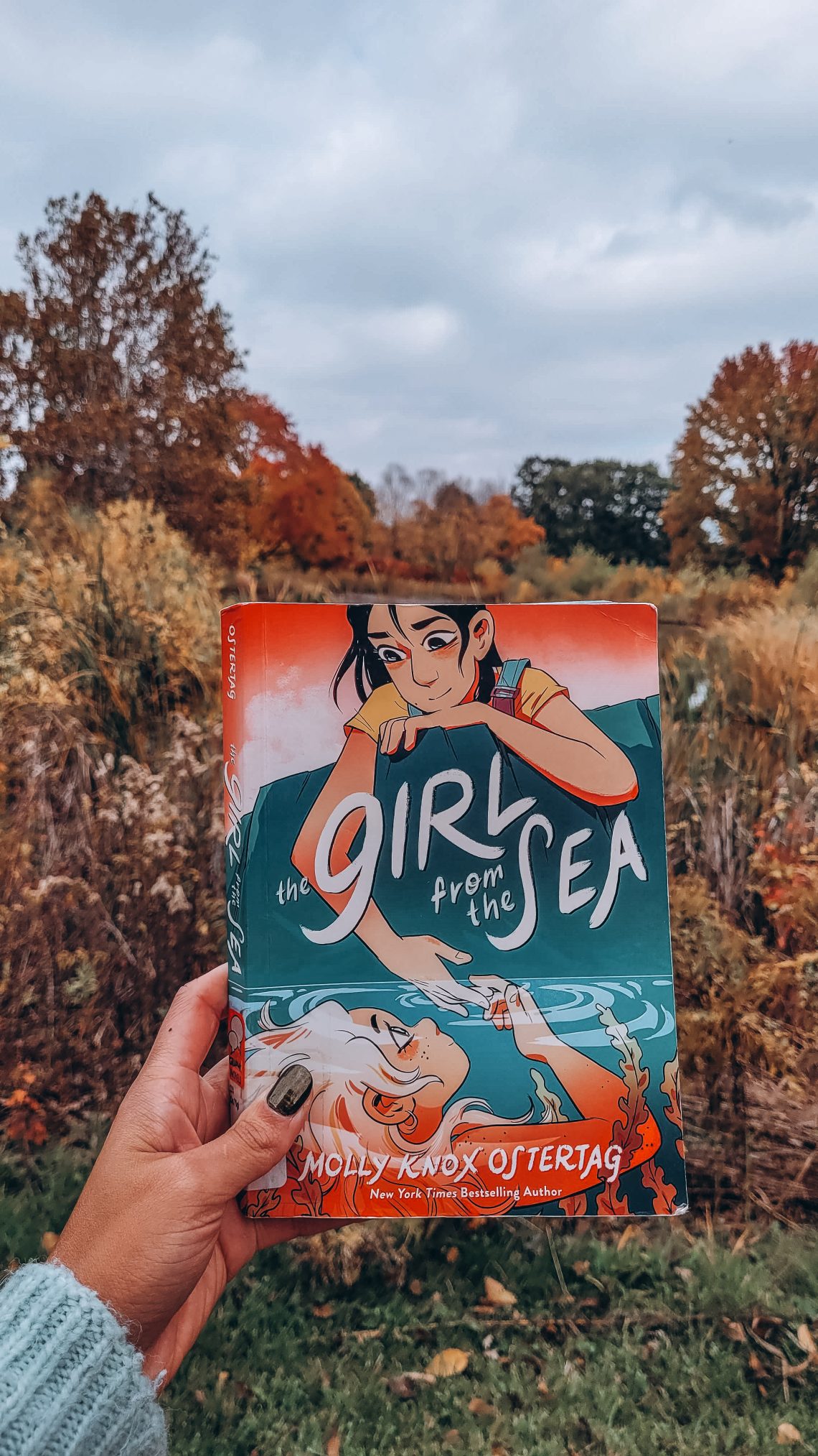 A light brown hand with dark nail polish holds The Girl from the Sea in front of a small lake with fall foliage