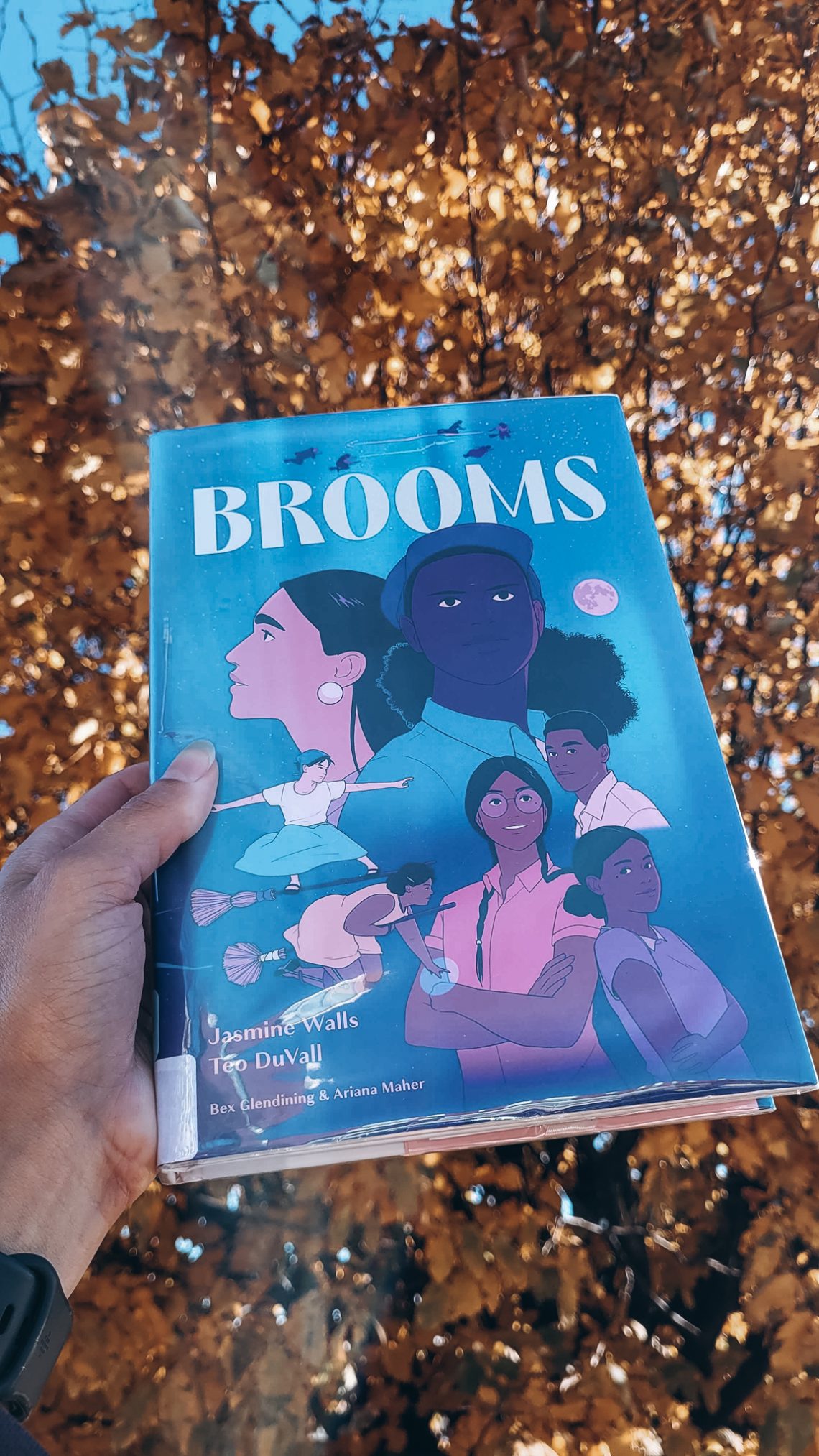 A library copy of Brooms held aloft by a light brown hand with golden leaves in the background